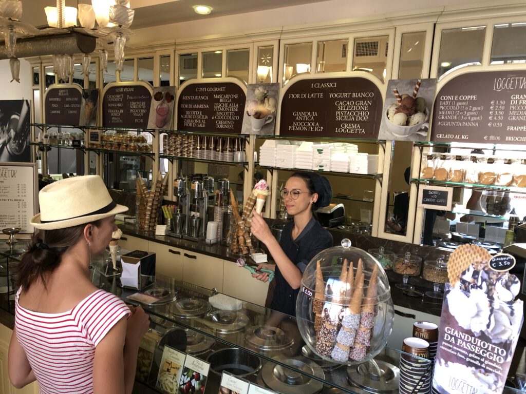 Eine Eisdiele (Gelateria) mit kunstvoll angerichtetem Gelato und einer Kundin davor. Perfekt für den Moment der Entschleunigung und des Genusses.